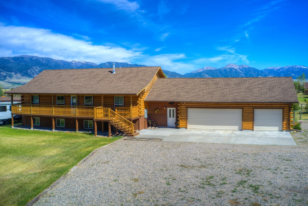 A spacious log cabin is presented against a backdrop of rolling mountains and a clear blue sky. The front porch features a wooden railing, and the garage doors are visible along the lower level. Gravel landscaping surrounds the home, offering ample outdoor space.