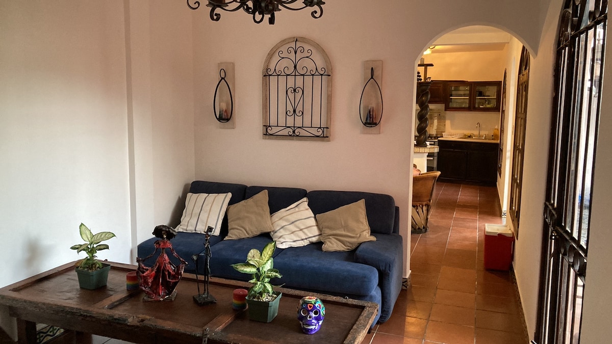 A comfortable sitting area features a blue couch adorned with striped cushions beside a rustic wooden table. Wall art and small potted plants add character. An archway leads to a well-equipped kitchen visible in the background, enhancing the inviting ambiance.