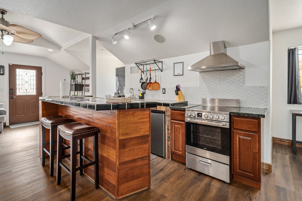 The kitchen area features modern appliances, including a stainless steel stove and refrigerator. A wooden breakfast bar with two stools offers seating. Wall-mounted shelves display kitchen essentials. The entrance door is visible, with natural light illuminating the open space.