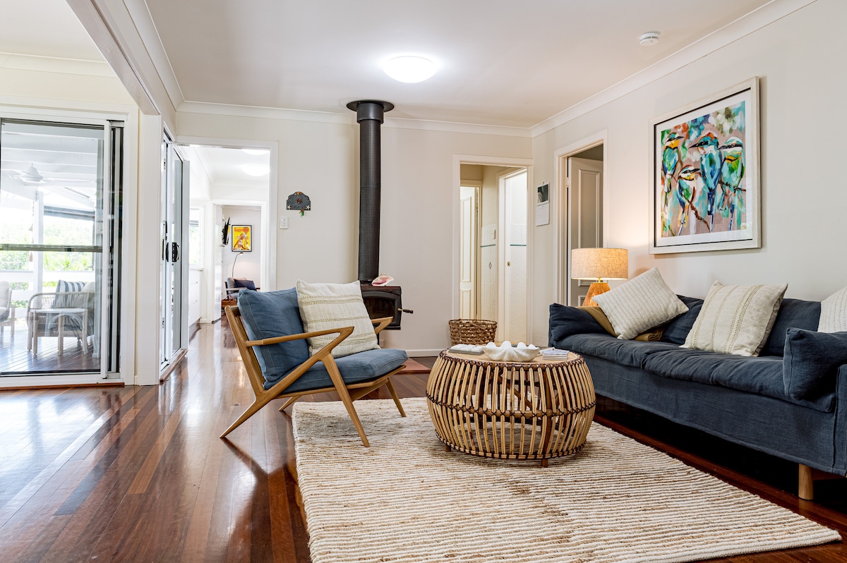 The living area features comfortable seating arranged around a circular coffee table. A rug covers the wooden floor, and a painting of birds is displayed on the wall. Glass doors provide views to the outdoor space, enhancing natural light in the room.