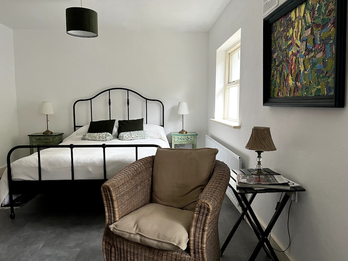 A bright and airy suite is showcased with a spacious bed framed in black metal. Two bedside lamps provide soft lighting. A comfortable wicker chair is positioned beside a small table, while a colorful piece of artwork adds a vibrant touch to the white walls.