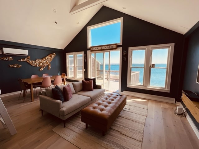 A spacious living area features large windows framing panoramic views of the harbor. Natural light floods the room, highlighting a brown leather sectional sofa and a wooden dining table. A decorative wall art element in the shape of a whale adds character to the inviting space.