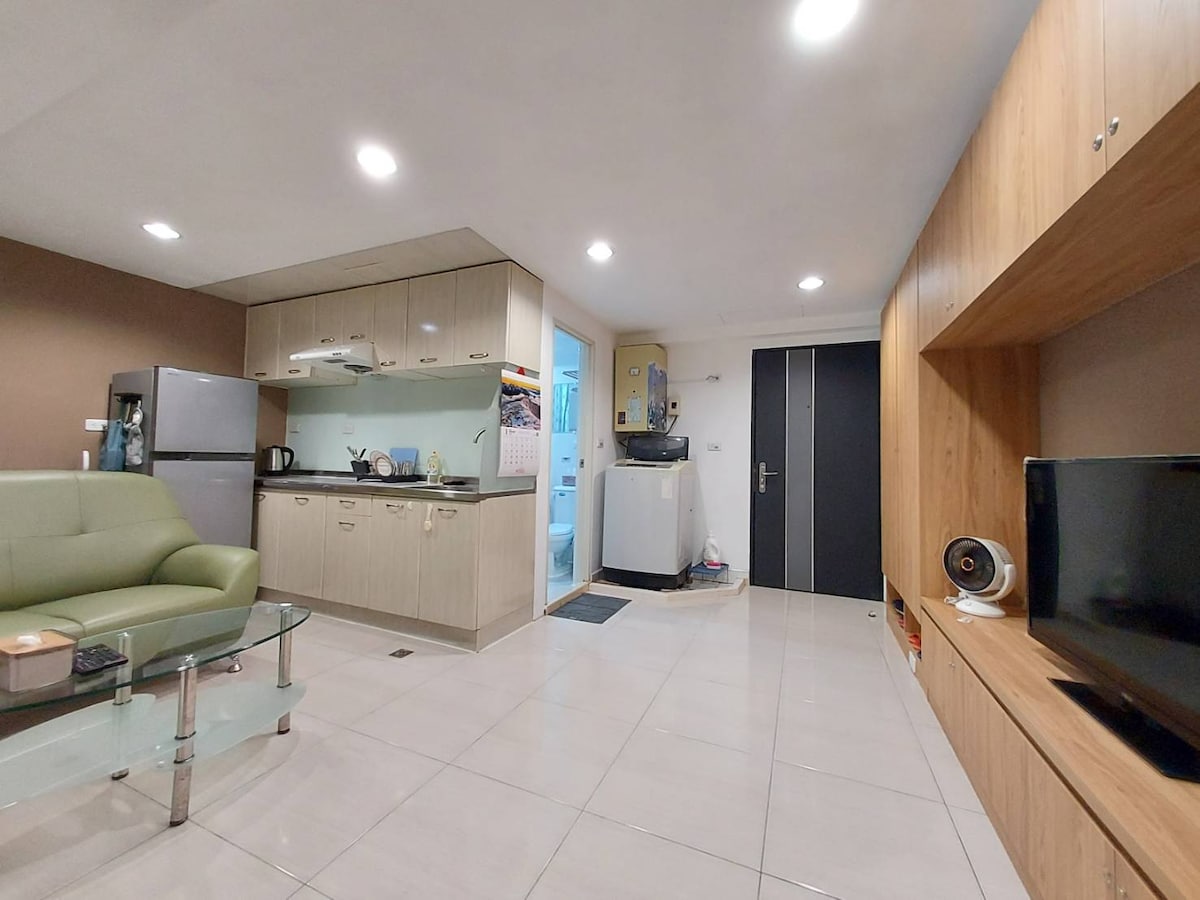 A modern living area is displayed with light-colored tiled flooring. The space features a green couch, a glass coffee table, and a wooden entertainment center housing a TV. A kitchenette with cabinets and appliances is seen, along with a washing machine in a corner.