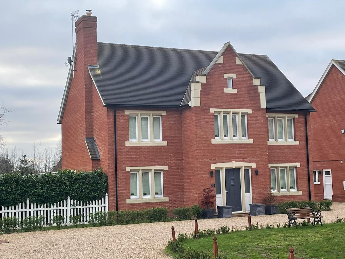 A well-maintained two-story brick house is surrounded by a gravel pathway and manicured hedges. Features include large windows that allow for ample natural light and a stylish front door, contributing to its modern yet classic appearance. A shared landscaped garden is visible in the background.