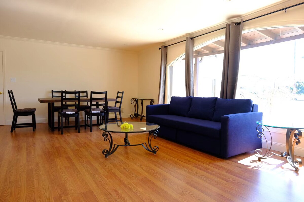 A spacious living area showcases modern furnishings, featuring a blue sofa, a round glass coffee table, and a dining table with six chairs. Natural light floods the room through large windows, illuminating the wooden floor and neutral-colored walls.