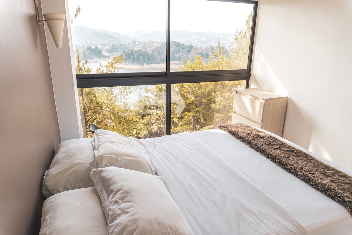 A modern bedroom features a large window revealing views of the natural landscape and nearby water. A spacious bed with white linens and soft pillows is complemented by a textured throw blanket. Minimalist wooden furniture enhances the serene atmosphere.
