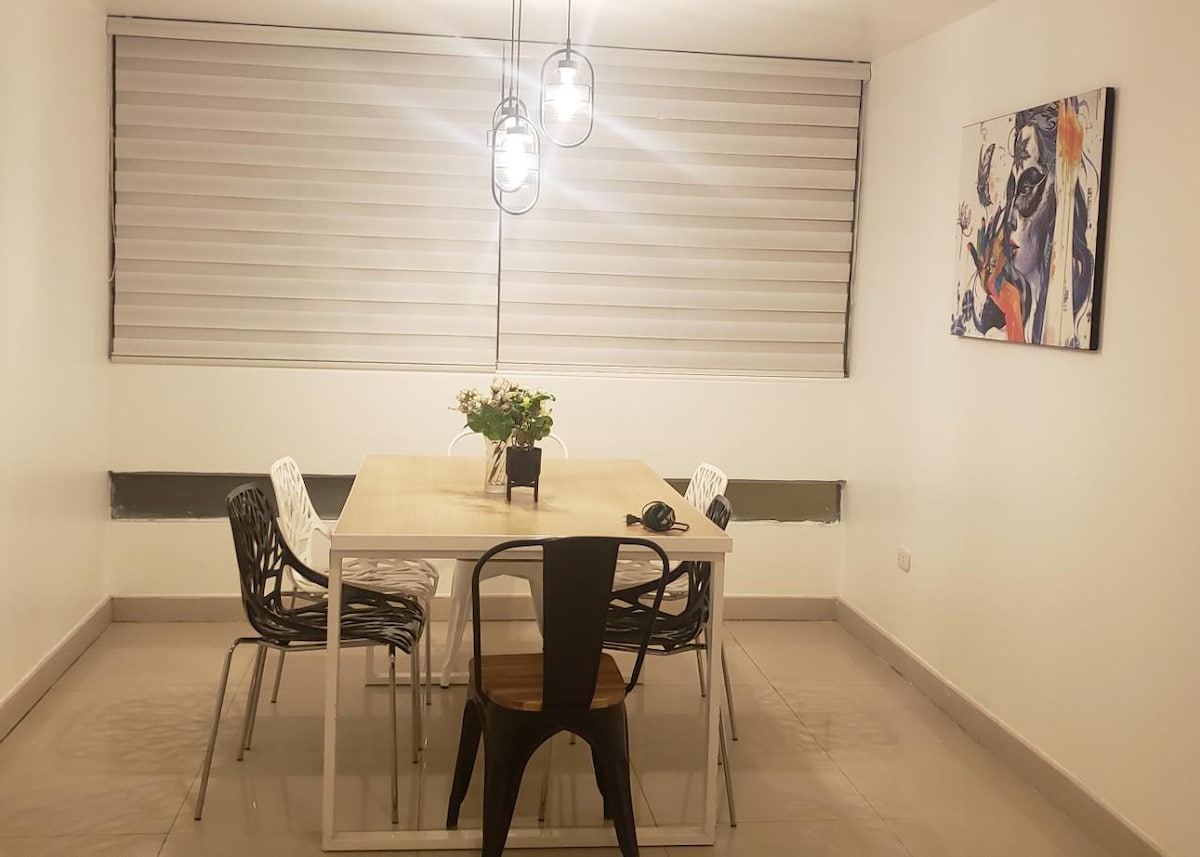 A dining area features a light wooden table surrounded by six black and white chairs. The space is complemented by pendant lighting above the table and a colorful wall art piece. Natural light is filtered through sleek window shades.