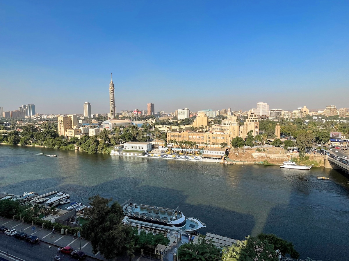 A panoramic view of the Nile River and Cairo cityscape is presented, showcasing modern buildings and historical architecture along the riverbank. The clear blue sky enhances the vibrant scene, with boats gently floating on the water's surface.