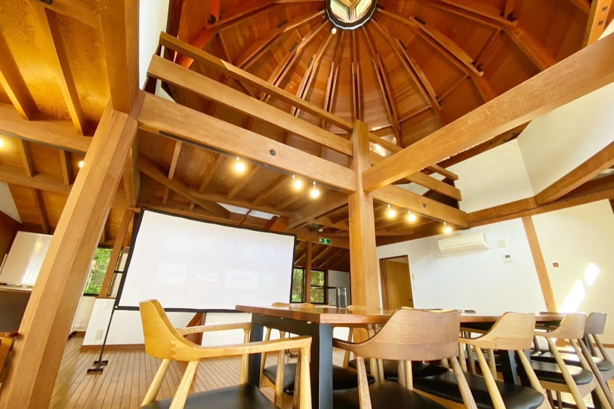 The interior features a high wooden ceiling with exposed beams, creating an open and airy atmosphere. A large projector screen is mounted on a wall, complemented by wooden furniture including a spacious dining table and several chairs. Natural light floods in, enhancing the warm wood tones.