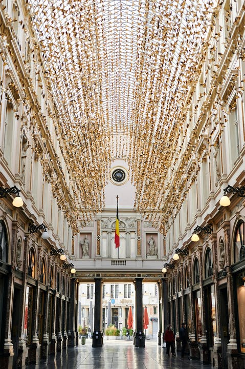 The image depicts a grand arcade with elegant architecture. A high, vaulted ceiling is adorned with numerous golden decorations, creating a striking visual effect. Soft lighting from hanging lamps casts a warm glow over the polished floor, while sculptures and windows can be seen along the walls.