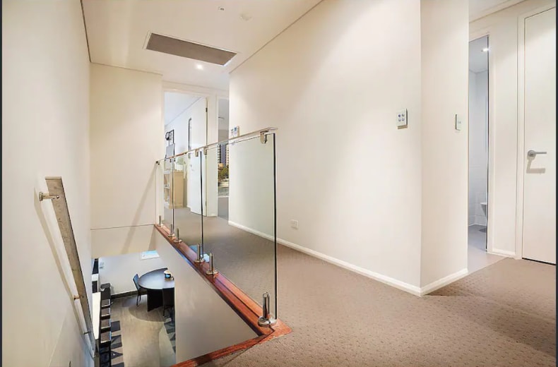 A spacious hallway is presented, featuring a modern glass railing that overlooks the lower level. Neutral-colored walls and carpeting create a light ambiance, while doors lead to various rooms. Soft lighting contributes to a welcoming atmosphere, enhancing the flow of the open-concept design.