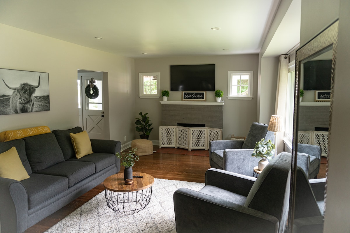 A welcoming living area features a comfortable seating arrangement with two gray sofas and two armchairs. A round coffee table sits at the center on a soft area rug. Natural light filters through the windows, illuminating the space, which includes a television above a low fireplace.