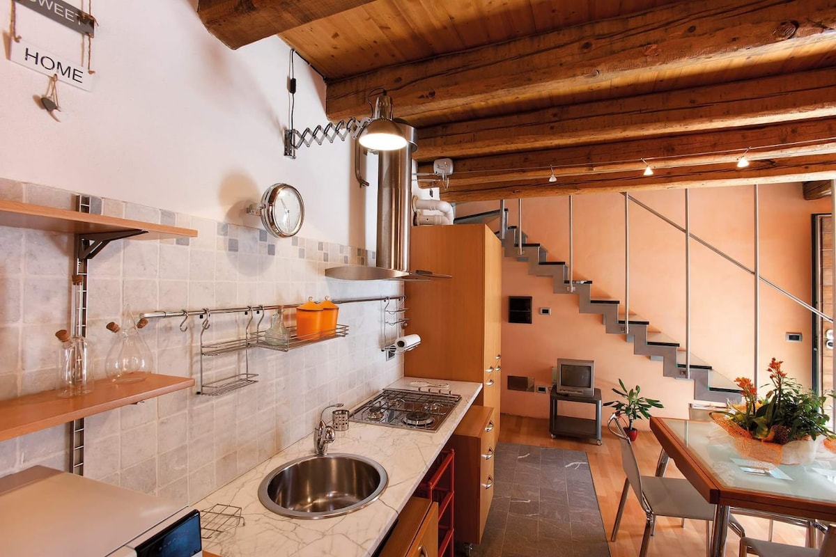 The kitchen area features a modern design with a gas stove, stainless steel sink, and wooden cabinetry. Open shelving displays glassware, while the space is complemented by rustic wooden beams overhead. A dining table with chairs is visible, alongside a staircase leading to the upper floors.