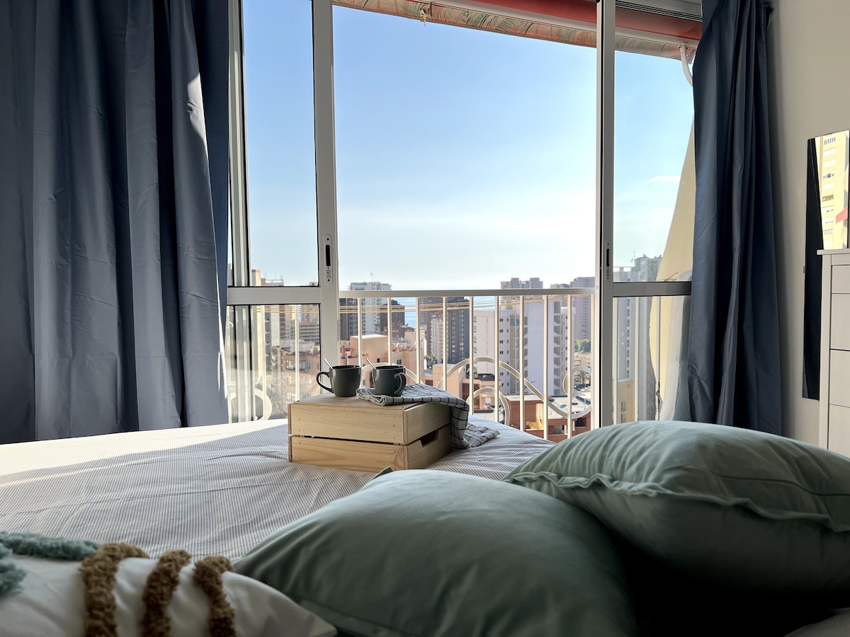 A bright sleeping area is illuminated by natural light streaming through large glass doors. A view of the sea and cityscape is visible in the background, while a tray with two cups sits on the bed, complemented by soft green pillows.