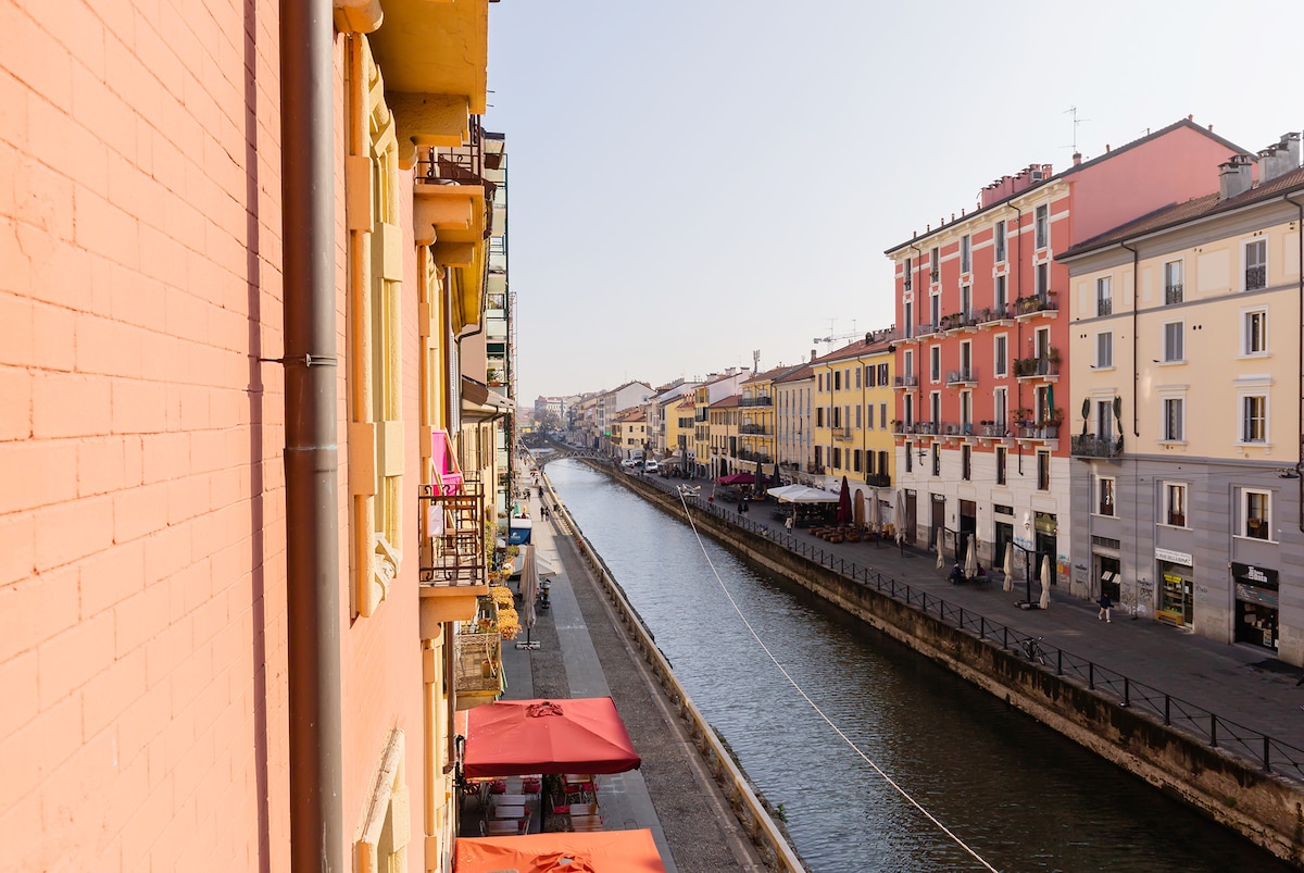 A view of the Naviglio Grande is displayed, with charming buildings lining the canal. Terracotta and pastel colors create a warm palette. Outside seating under umbrellas is visible along the waterway, inviting leisurely moments. The scene captures the essence of the vibrant neighborhood.