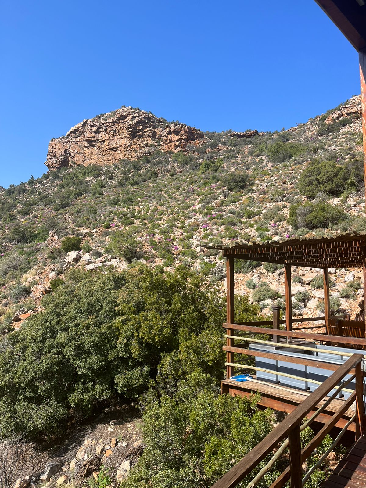A sweeping view of natural landscapes is captured from the veranda. Lush greenery covers rolling hills, with rugged rock formations rising prominently in the background under a clear blue sky.