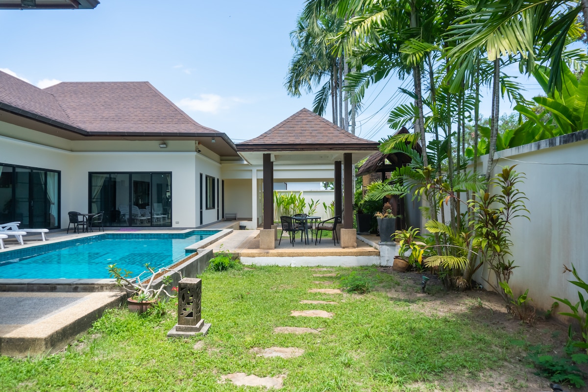 A private outdoor space features a clear swimming pool surrounded by a well-manicured garden. A gazebo with a dining table and chairs is positioned near the pool, and tropical plants line the perimeter, adding greenery to the tranquil environment.