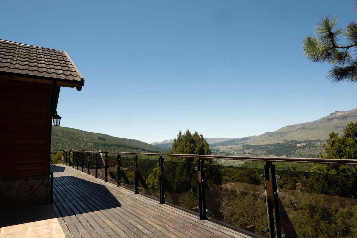 A wooden deck extends from the house, offering unobstructed views of the surrounding valley and mountains. The clear sky creates a serene backdrop, while glass railings enhance the openness of the space.