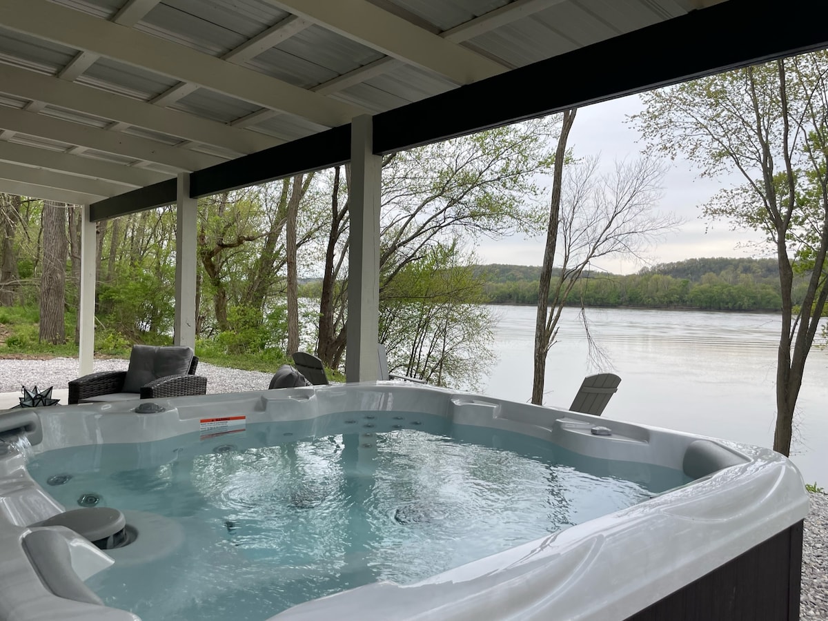 A six-person hot tub is positioned on a covered patio with a view of the river. The water flows gently, reflecting the surrounding trees and landscape. Relaxing seating options are visible nearby, inviting guests to enjoy the serene atmosphere by the water.
