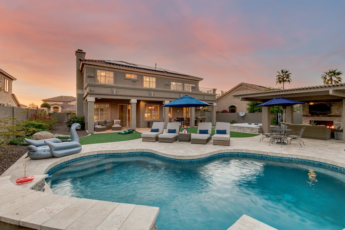 A spacious backyard features a shimmering pool with a water feature, surrounded by lounge chairs and an inflatable pool float. Umbrellas provide shade over the lounging area. The patio includes a dining set and a cozy seating area beside a fireplace, set against a pastel sunset sky.