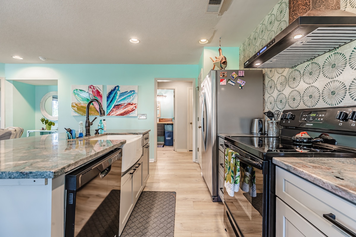 The kitchen features a central island with a sink and contemporary appliances. Light-colored cabinetry complements the spacious countertops. A colorful wall art piece adds a lively touch, while natural light filters through a nearby window, illuminating the modern design.