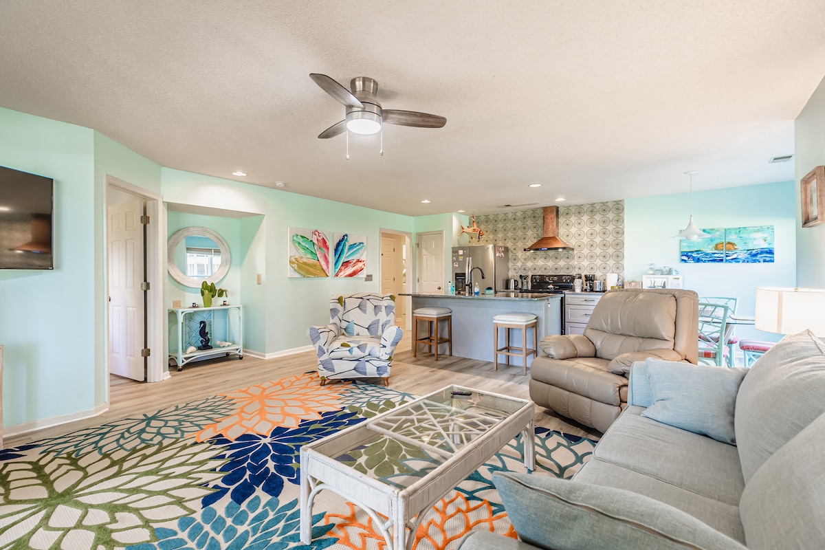 A spacious living area features a light color palette with aqua green walls and decorative wall art. Comfortable seating includes a patterned armchair and a plush sofa. A coffee table is centered on a vibrant area rug, while a modern ceiling fan enhances airflow in the room.