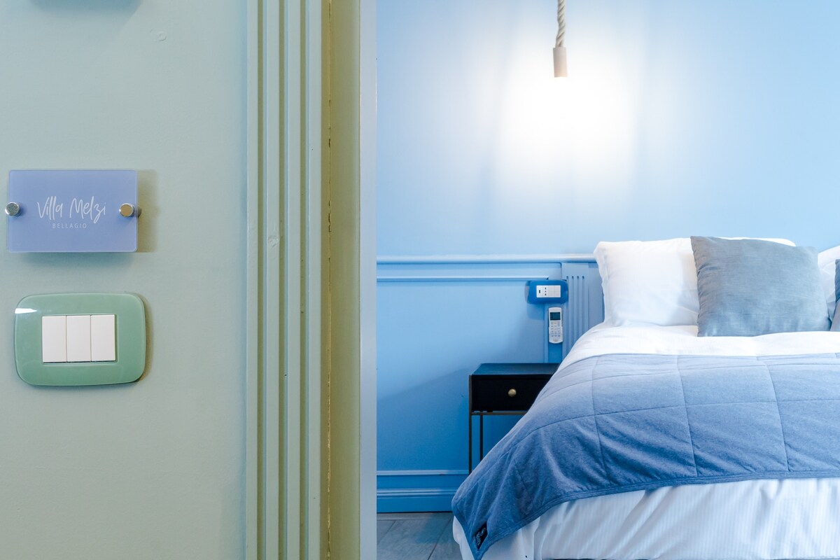 A serene bedroom is visible, featuring soft blue walls and a spacious bed dressed in light linens. A bedside table is positioned next to the bed, and a light fixture illuminates the area, creating a calming ambiance. The entrance door with a room sign is also in view.