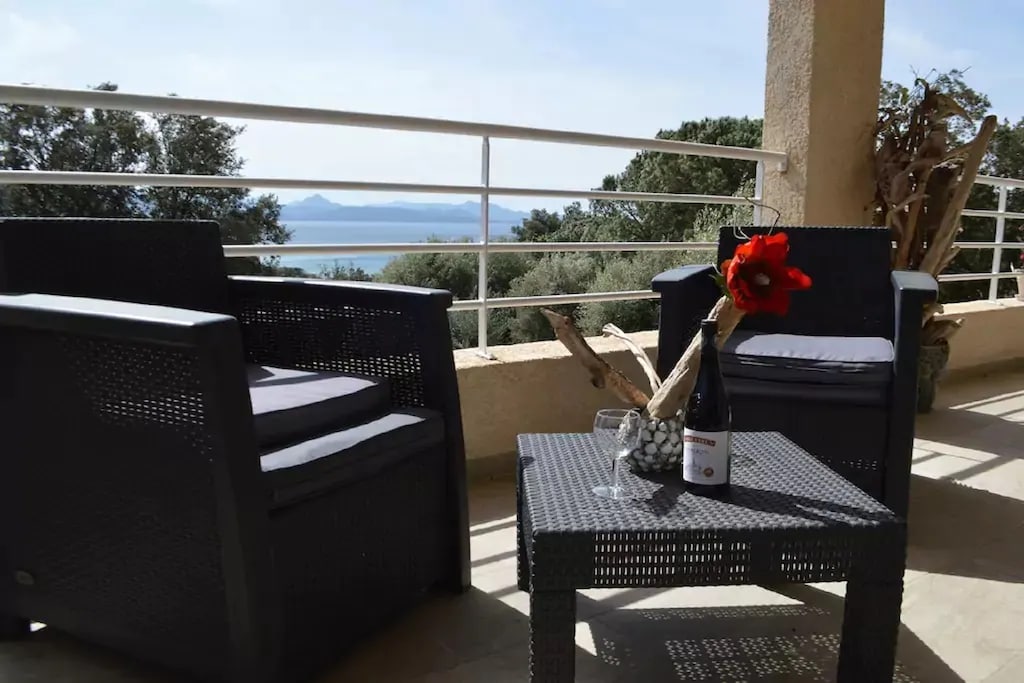 Two outdoor chairs are positioned around a small table, displaying a decorative flower and a wine bottle accompanied by glasses. The surrounding area features a railing with a serene view of the sea and greenery in the background.