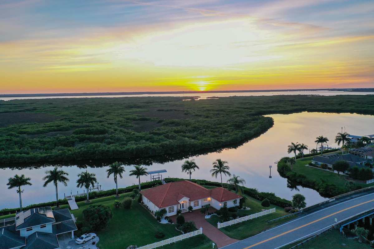 Aerial view of a waterfront property featuring a modern single-story house with a red-tiled roof. Surrounding palm trees are visible, and the serene waterway reflects the sunset colors. Lush greenery is present nearby, enhancing the peaceful ambiance of the landscape.