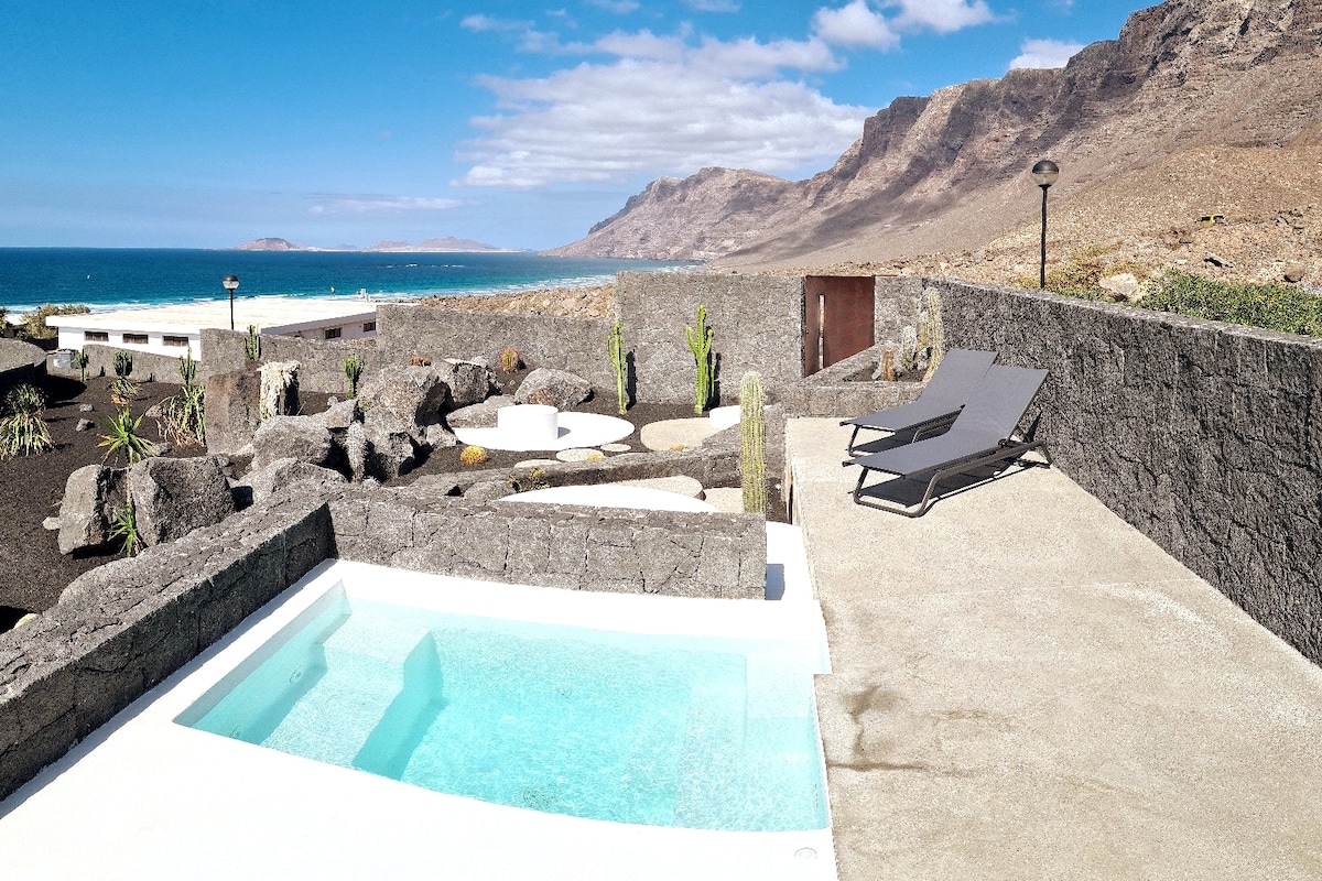 A small private pool with clear water is surrounded by smooth concrete and natural stone elements. Two lounge chairs are positioned nearby, offering unobstructed views of the ocean and mountains. The sky displays a mix of clouds and sunshine, enhancing the coastal scenery.