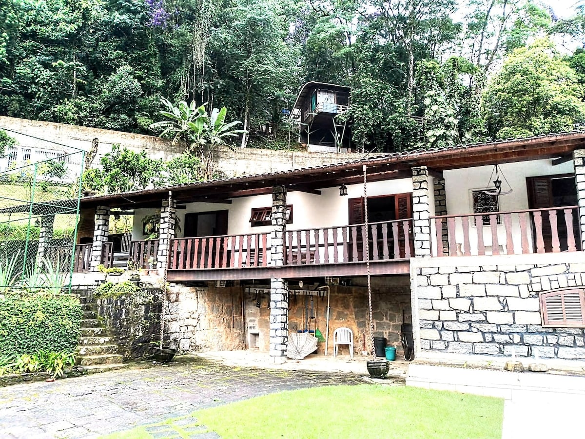 A charming house is nestled within lush greenery, showcasing a spacious covered porch supported by stone columns. Steps lead to a garden area, while trees and plants surround the property, enhancing the natural ambiance. A second structure is visible in the background among the trees.