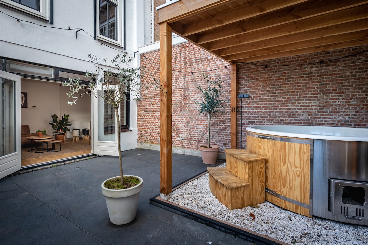 A spacious patio is surrounded by brick walls, featuring a wooden structure supporting a round jacuzzi. Decorative olive trees are positioned in planters, while a gravel bed enhances the outdoor space. Large glass doors provide access to the interior living area.
