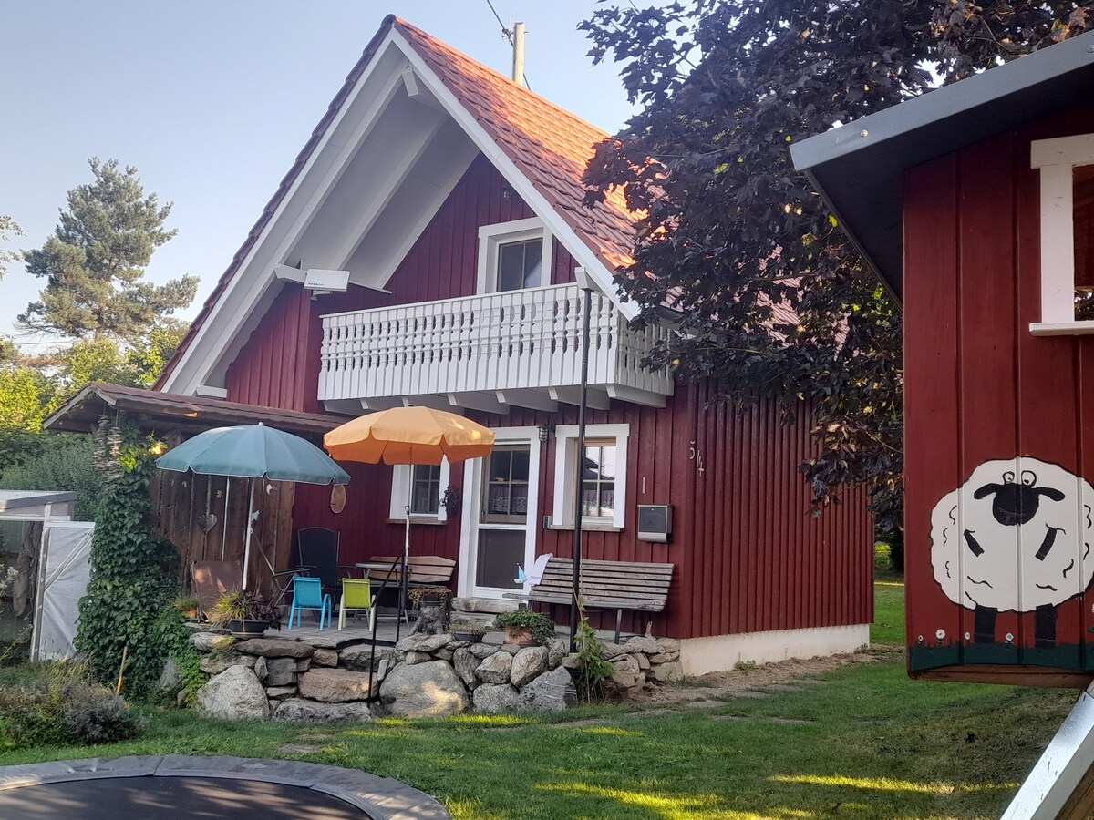A charming red wooden house is surrounded by greenery, featuring a balcony and outdoor seating under umbrellas. A stone patio area with chairs invites relaxation, while a trampoline is visible in the foreground, adding a playful element to the landscape.