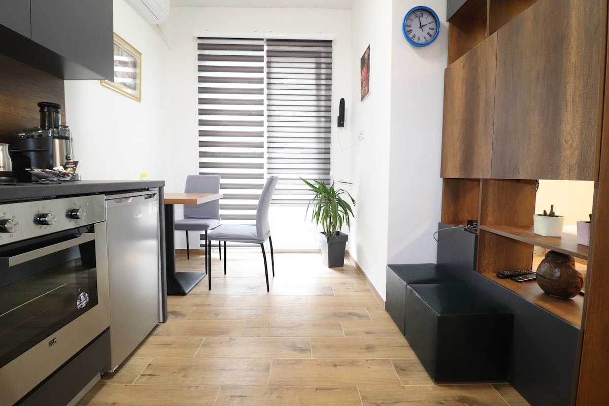 A small kitchen area features modern appliances and cabinetry. A dining table with two chairs is positioned next to a large window, adorned with striped blinds, allowing natural light to enter. A decorative plant and a clock are placed along the wall, enhancing the atmosphere.