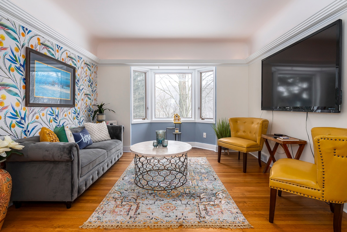 A bright living room features a comfortable gray sofa and two yellow chairs positioned around a decorative coffee table. Large windows allow natural light to fill the space, complemented by colorful wallpaper displaying floral patterns. A mounted TV is located on the wall opposite the seating area.