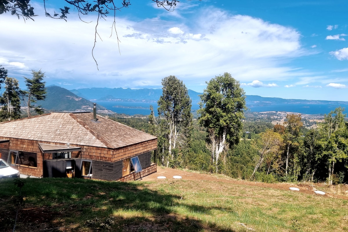 A cozy house with a wooden shingle roof sits on a hillside, surrounded by trees. The expansive view reveals Lake Ranco and snow-capped mountains in the distance, under a bright blue sky with soft clouds. A grassy area in the foreground complements the landscape.