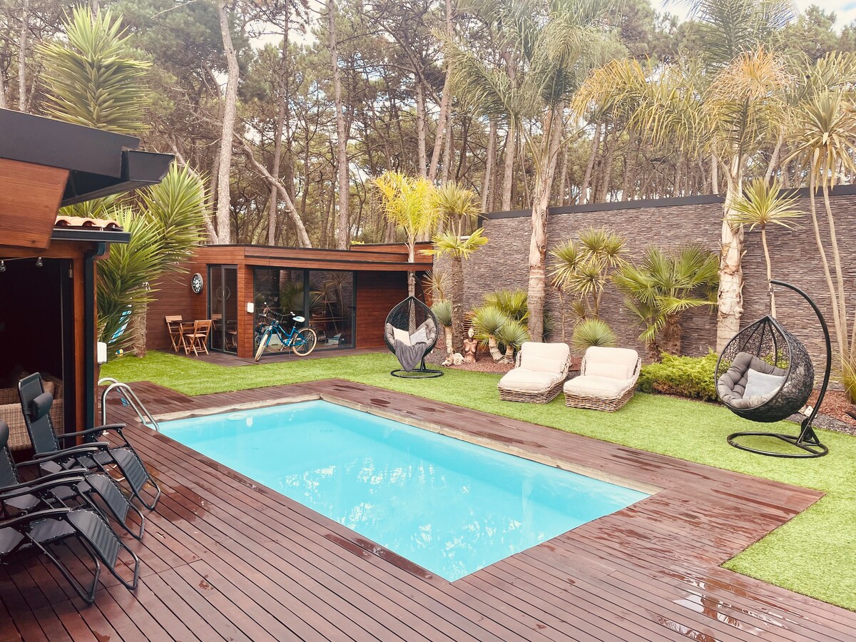 An outdoor space features a rectangular pool surrounded by a wooden deck and well-maintained greenery. Two hanging chairs provide seating, while two lounge chairs are positioned nearby. A bicycle can be seen resting against a wall, adding to the tranquil atmosphere.