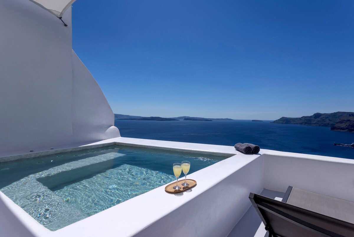 A private Jacuzzi hot tub is positioned on a sun-drenched terrace, offering expansive views over the azure caldera. Two glasses of sparkling beverage are placed on a wooden serving tray beside the tub, with a towel rolled neatly nearby. The clear sky enhances the serene atmosphere.