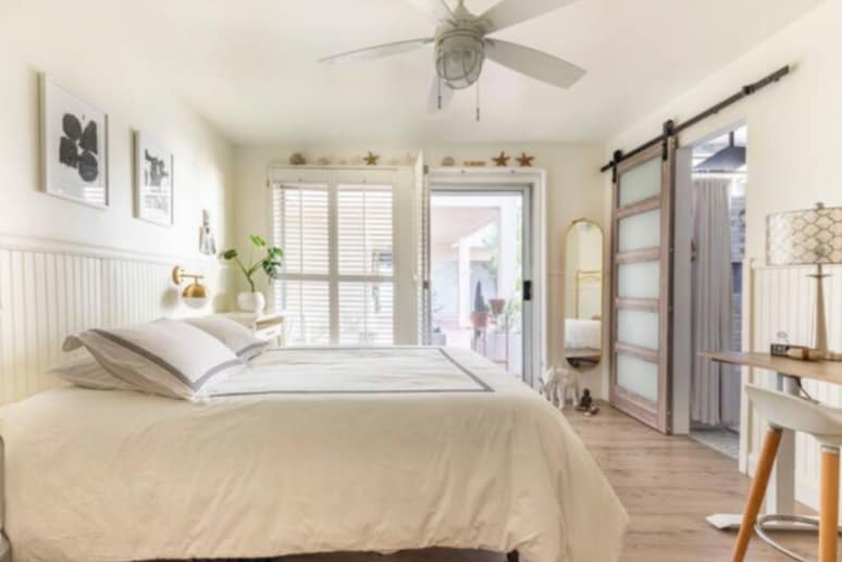 A well-lit bedroom features a spacious bed with crisp white bedding. Natural light enters through large windows adorned with wooden slats. A fan hangs from the ceiling, while a sliding door provides access to an outdoor space. Decorative elements include framed art and a floor lamp.
