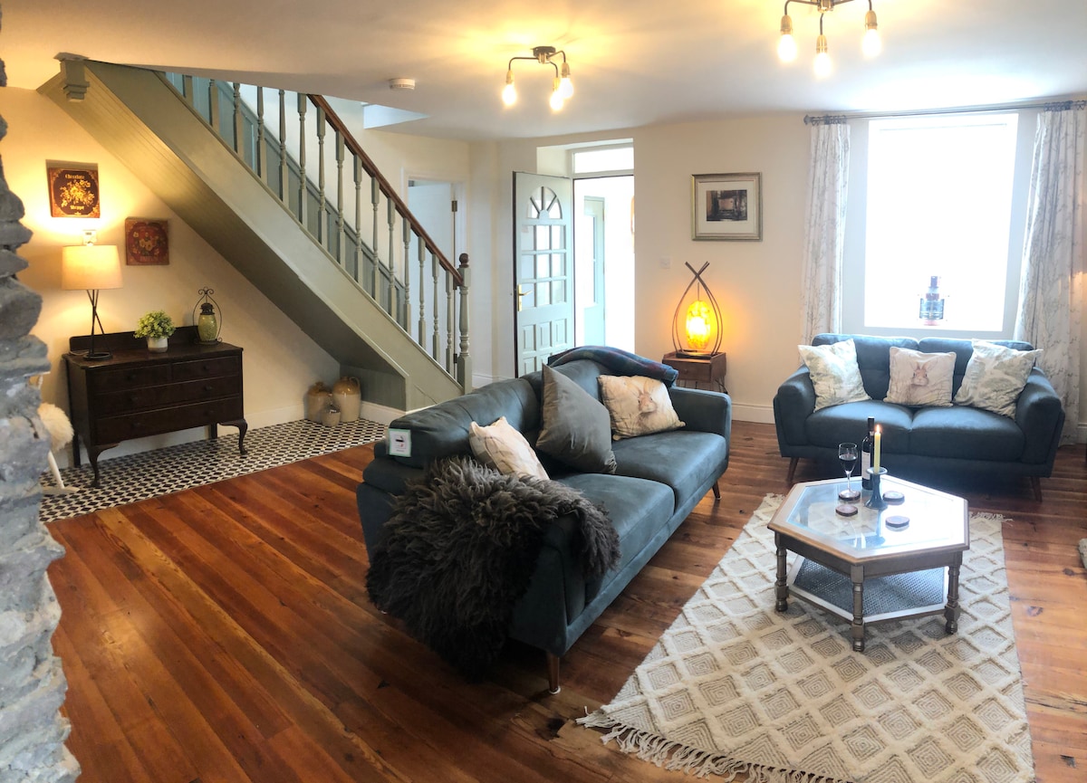The open-plan living area showcases original pitch pine flooring, with two comfortable couches positioned around a wooden coffee table. A textured area rug complements the space, while natural light floods in through the large windows. Staircase and entryway are visible in the background.