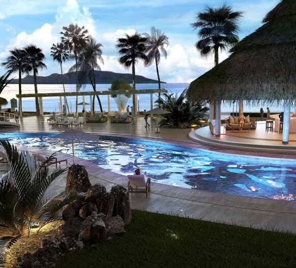 An inviting outdoor pool is surrounded by lush landscaping, including palm trees and decorative rocks. Lounge chairs are positioned around the pool area, with a thatched roof bar visible in the background. A serene ocean view complements the tranquil setting.