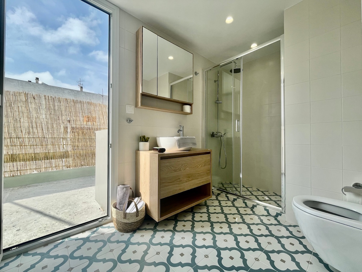 A modern bathroom features a spacious shower area enclosed in glass. A stylish wooden vanity is complemented by a large mirror. Decorative patterned tiles cover the floor, while natural light streams through a sliding glass door, leading to an outdoor space.