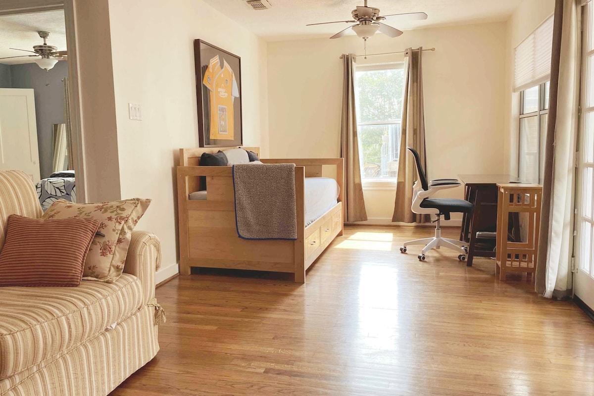 A bright bedroom area displays a daybed adorned with a blanket and a framed sports jersey on the wall. A desk with an office chair is positioned next to a window, allowing natural light to fill the space. Hardwood flooring enhances the room's openness.