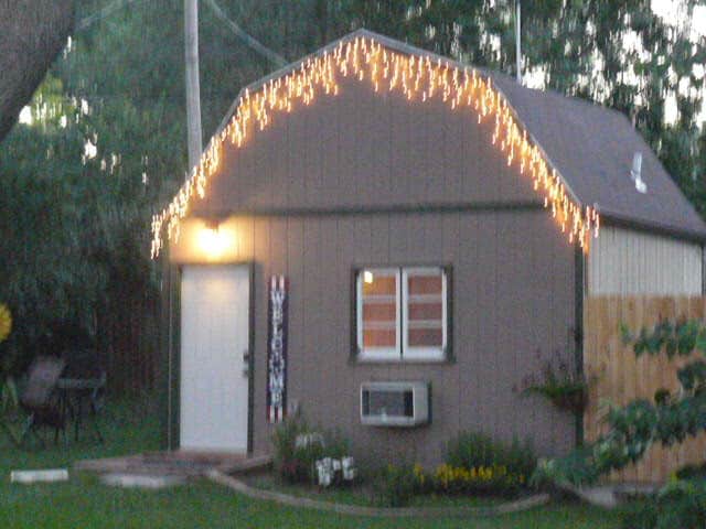 A cozy barn exterior is adorned with warm white string lights along the roofline. A welcoming entrance with a white door is visible, along with a few windows. The surrounding grassy area features flowering plants and a shaded sitting area with chairs.