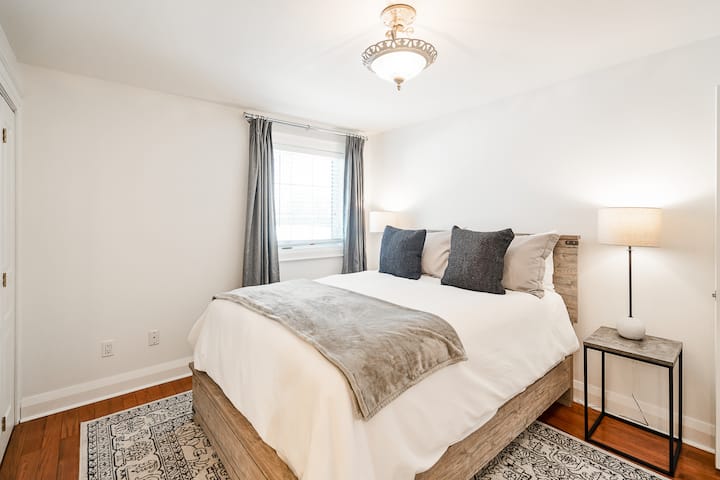 Main level queen bedroom with clean linens, closet, bedside lighting, window covering curtains, private ensuite bathroom. 