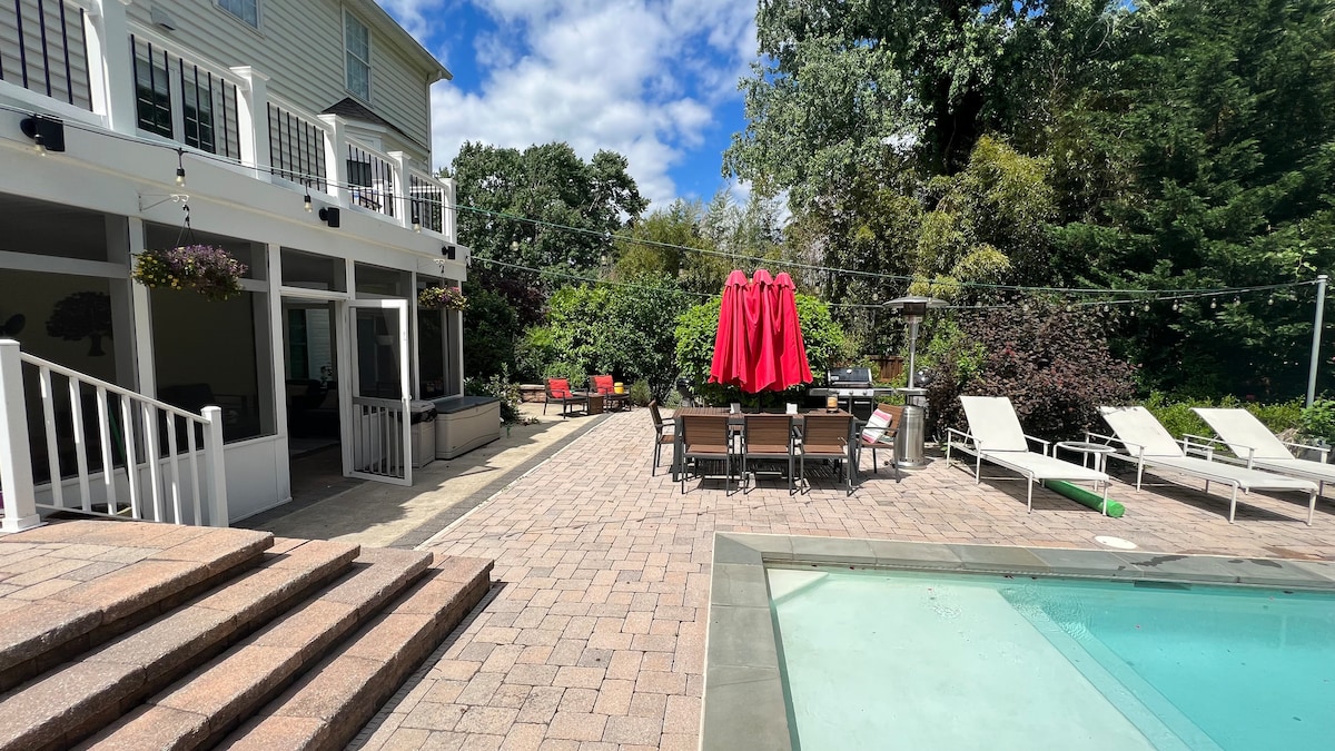 An outdoor space features a swimming pool bordered by lounge chairs and a dining area with a large table shaded by a vibrant red umbrella. Lush greenery surrounds the area, complemented by stone paving and steps leading to the entrance.