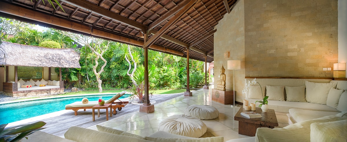 A spacious lounge area features soft, neutral furnishings that invite relaxation. Large open spaces are enhanced by natural light filtering through the wooden beams. The view overlooks a private pool surrounded by lush greenery, with a bale visible in the background, offering a serene outdoor space.