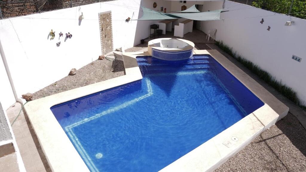 A refreshing swimming pool is surrounded by pale stone and white walls. A circular jacuzzi is positioned to one side, partially shaded by a canopy. Decorative elements are visible along the white wall, complementing the outdoor space.