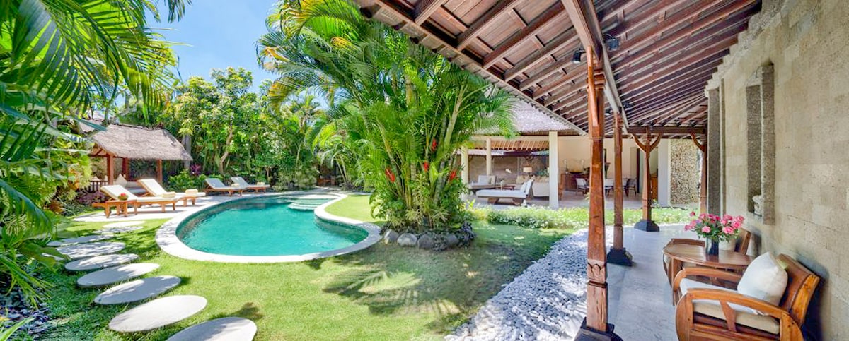 A private pool is framed by lush tropical landscaping, with lounge chairs positioned around its perimeter. A pathway of stepping stones leads to a shaded outdoor seating area adorned with decorative plants, creating a serene ambiance in the villa's garden.