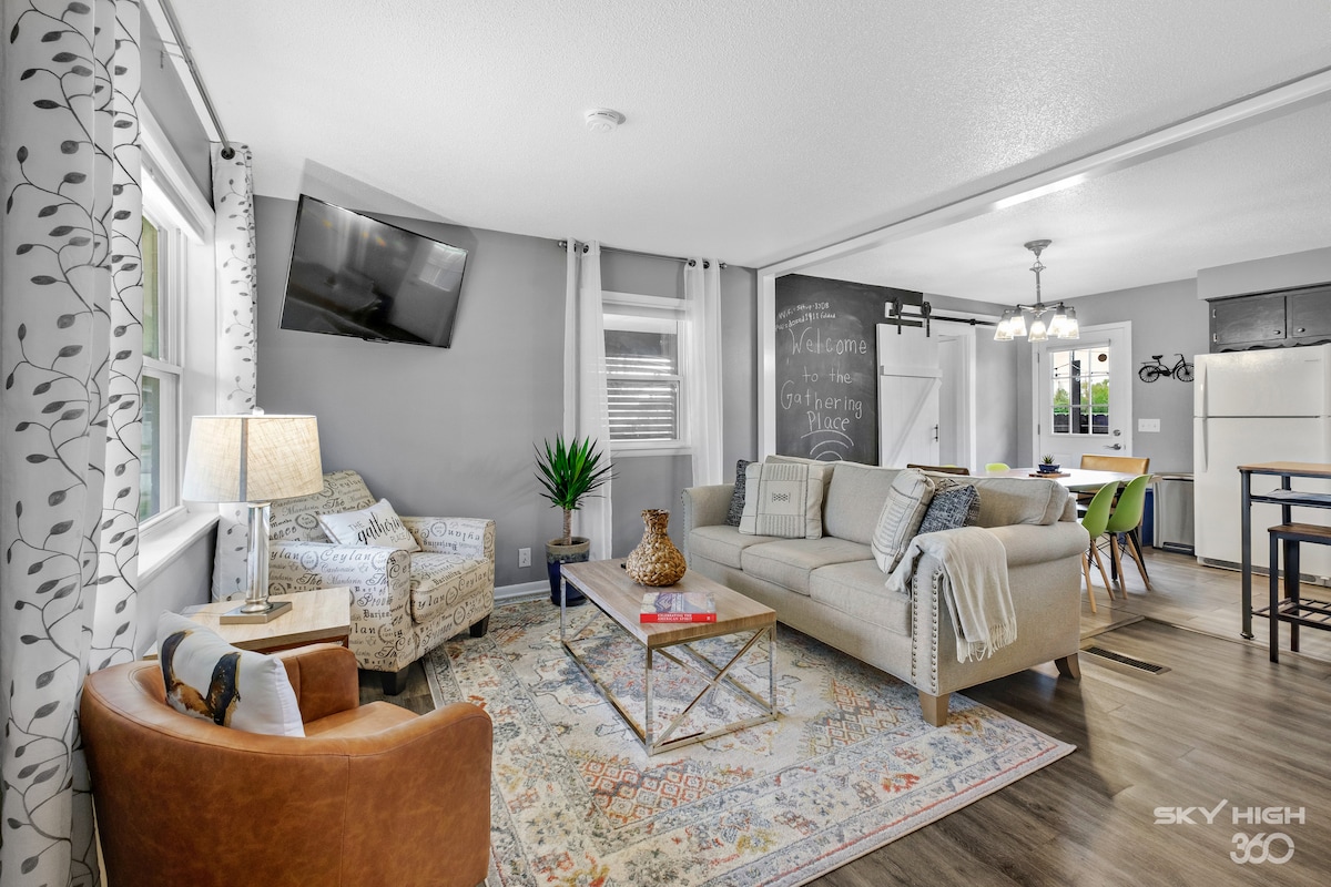 A bright living area features comfortable seating, including a beige sofa and a patterned armchair. A decorative area rug complements the space, while natural light streams through large windows. A television is mounted on the wall, and a chalkboard adds a playful element to the design.